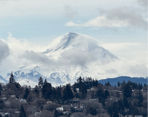 Snow-capped Mount Baker looms behind a town nestled among evergreen trees, showcasing a serene natural landscape under a cloudy sky.