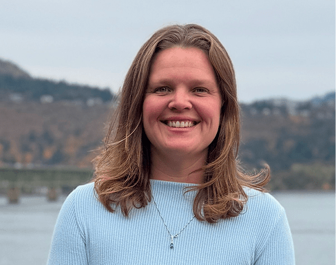 Smiling woman with shoulder-length brown hair stands by a river, wearing a light blue sweater. Soft landscape in the background adds a tranquil vibe.