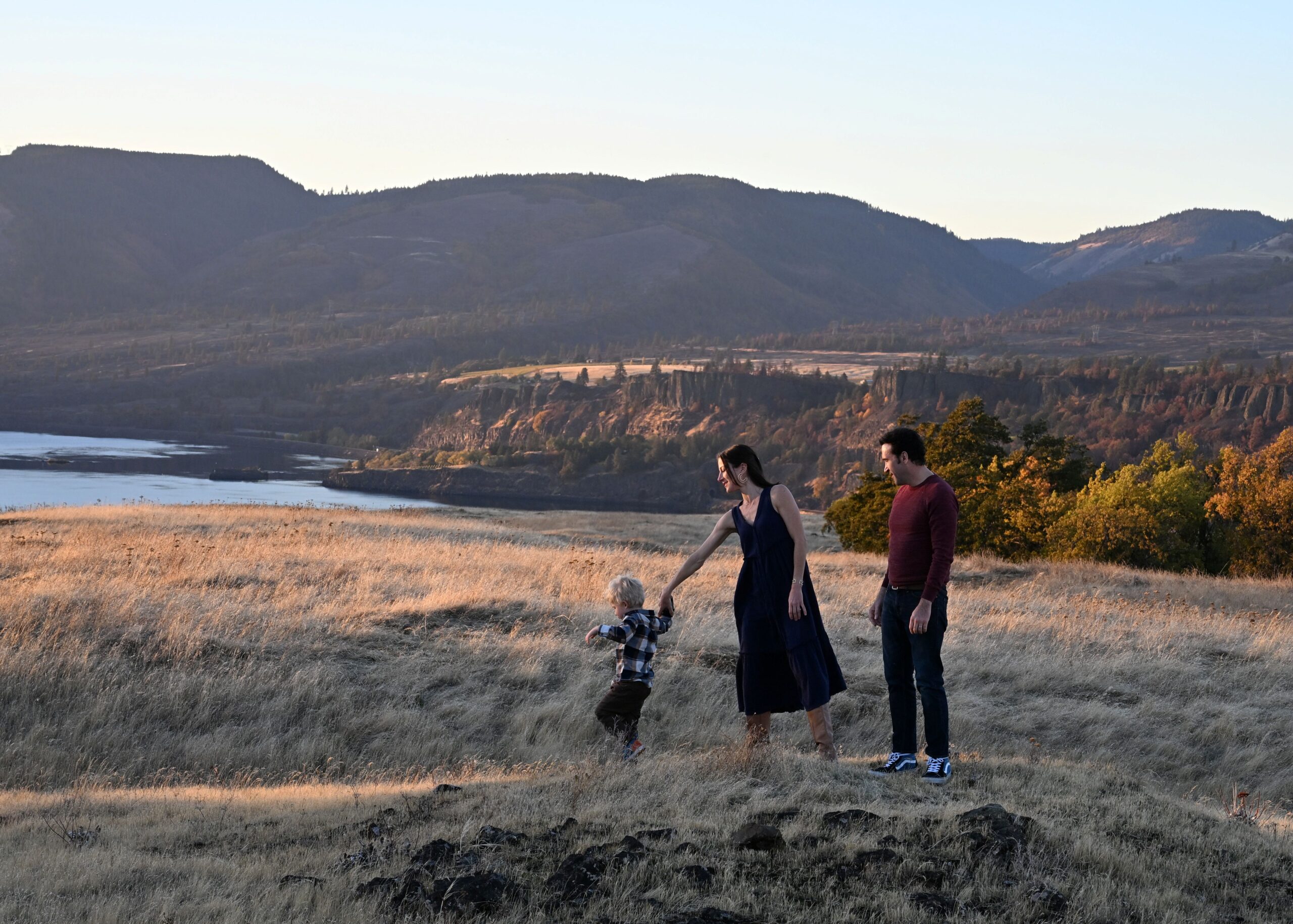 A woman gently guides a young boy while a man watches, surrounded by golden grass and a scenic landscape at sunset. The image highlights family bonding in nature.