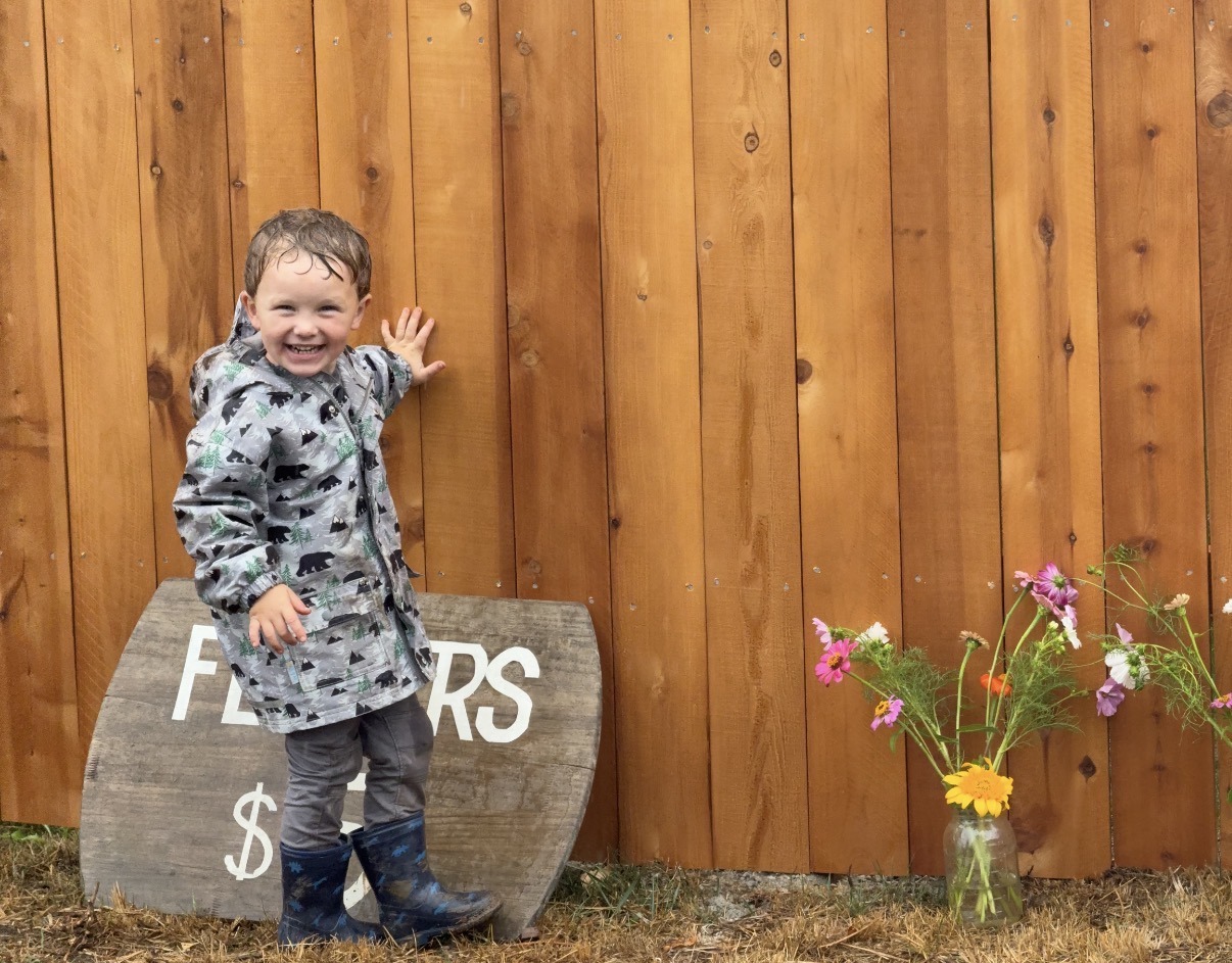 Smiling child in a raincoat poses by a wooden fence, next to a flower stand sign and a jar of colorful wildflowers, capturing a joyful moment amidst a rainy day.
