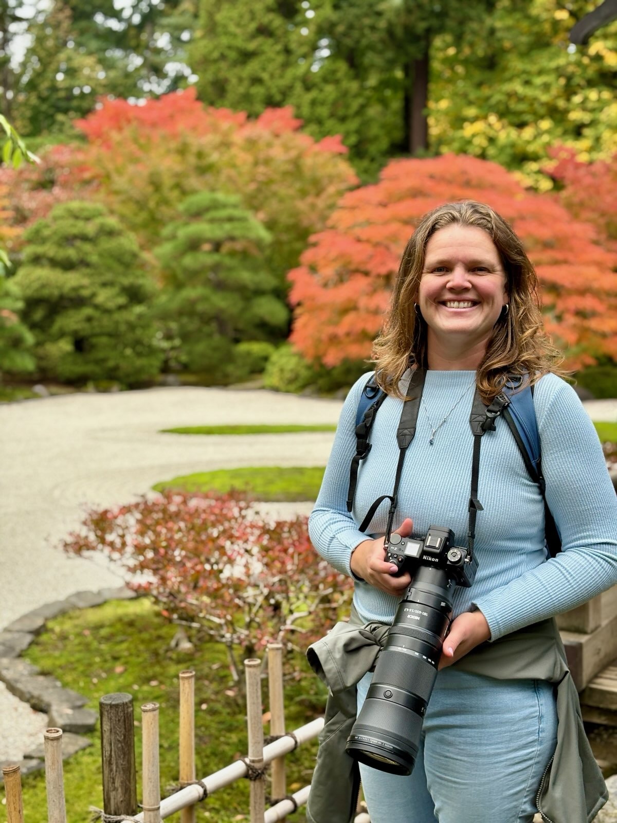 A smiling woman holds a camera in a vibrant garden, showcasing colorful autumn foliage, highlighting her passion for photography.