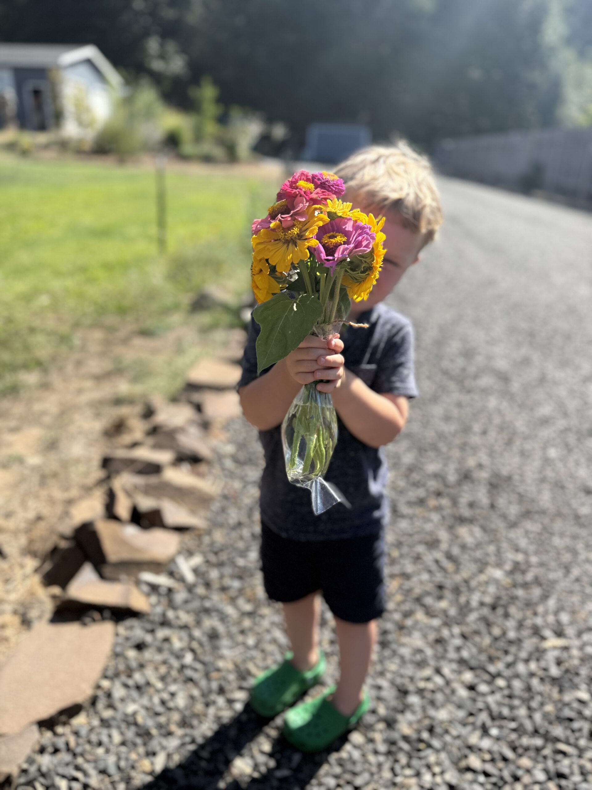 A young child with a short haircut holds a colorful bouquet of flowers in a glass vase. A gravel pathway and house are in the background.
