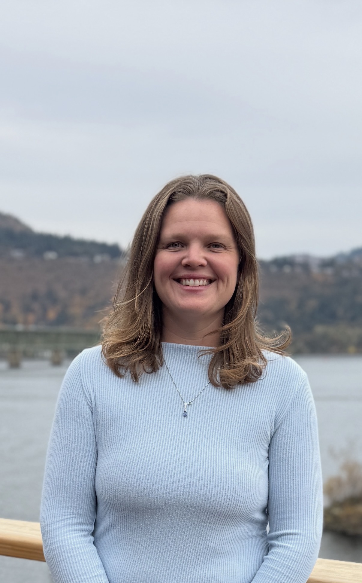 Smiling woman with long hair in a light blue ribbed sweater stands in front of a tranquil river and hills, emphasizing a serene outdoor setting.