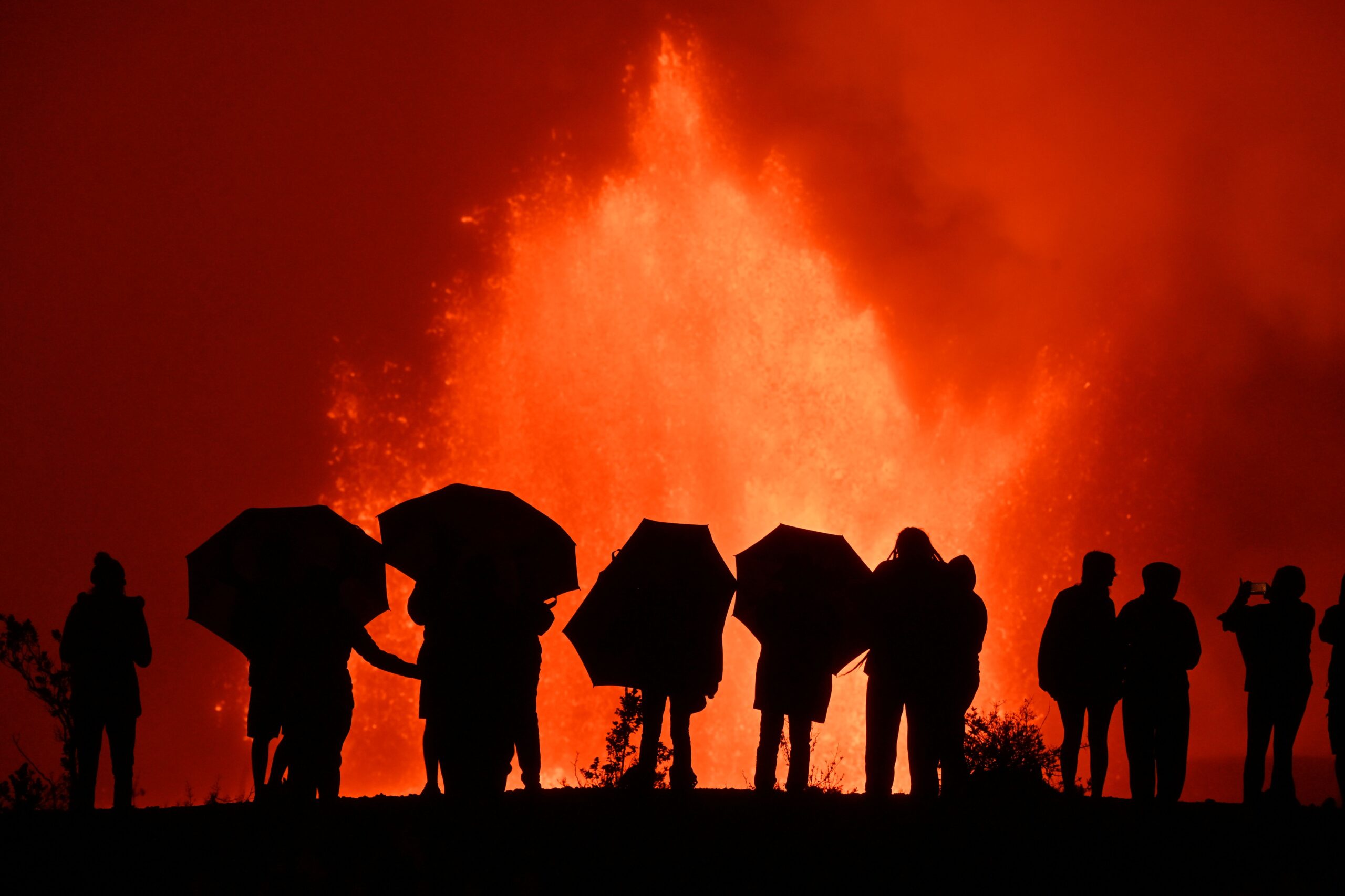 Silhouetted figures holding umbrellas watch a volcanic eruption, with molten lava spewing against a vivid red sky, showcasing nature's power.