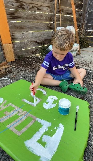 Young child painting a green sign with white letters on a stone surface, showcasing creativity and outdoor crafting.